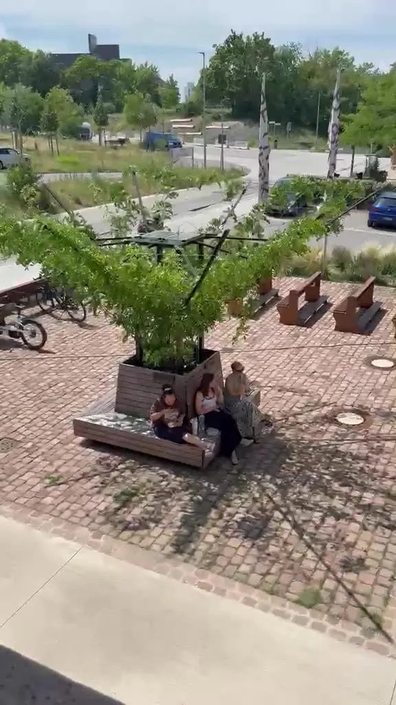 Film, Aufnahme vor Ort im Juli 2026: ausgeführte Sonnenarkade auf einem Platz, Menschen sitzen im Schatten unter dem begrünten Dach